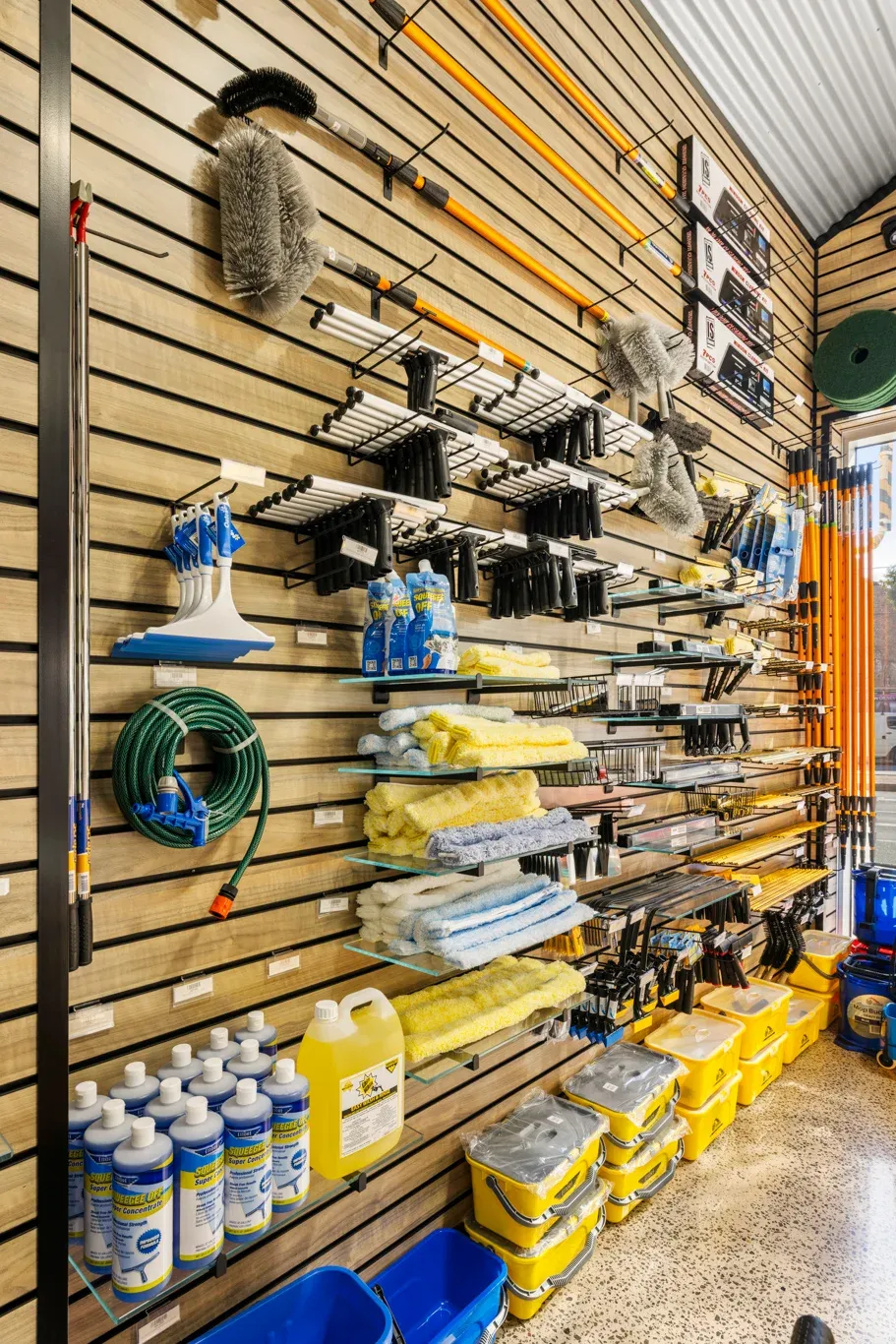 Trade showroom wall display featuring walnut slatwall panels with black inserts and long slatwall hooks for hanging cleaning tools, brushes, mop handles, and accessories.